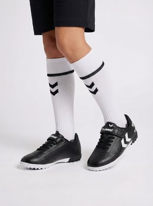 Beine mit weißen knielangen Socken mit schwarzen Chevron-Mustern und schwarzen Sportschuhen mit weißen Sohlen und hummel-Logo.