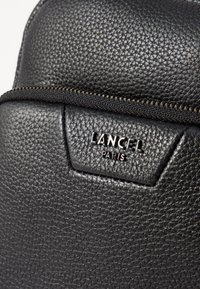 Détail d'un sac à dos en cuir noir, avec une finition texturée et un logo métallisé "LANCEL PARIS" sur un patch triangulaire. Fermeture éclair visible.