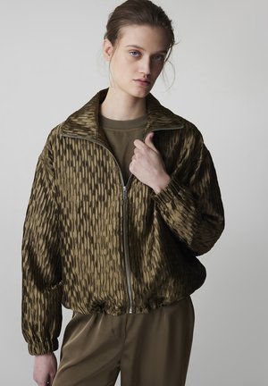 Junge Frau in olivgrüner, strukturierter Reißverschlussjacke mit hohem Kragen, passendem Hemd und Hose, stehend vor hellem, einfarbigem Hintergrund.