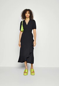 Black wrap dress with short puff sleeves, ruffled hemline, paired with bright green chunky platform sandals and a small green handbag.