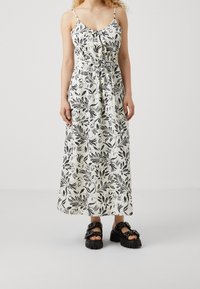Robe maxi avec un motif de feuilles noires sur tissu blanc, dotée de bretelles spaghetti ajustables et d'une taille cintrée par un cordon. Portée avec des sandales noires épaisses.