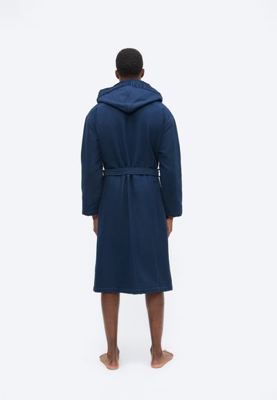 Άνδρας που στέκεται ξυπόλυτος με την πλάτη, φορώντας μια μπλε-σκούρα μπανιερό ρόμπα μέχρι το γόνατο με υφή, κουκούλα και ζώνη δεμένη στη μέση.