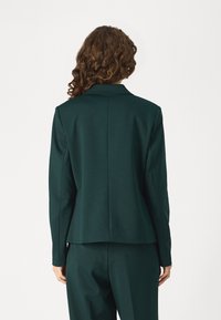 Veste blazer vert foncé avec une coupe ajustée, des revers crantés et une texture lisse. Elle possède une fente au dos et des épaules structurées.