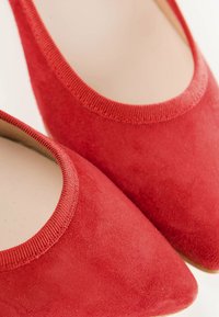 Los zapatos planos de punta ovalada en ante rojo presentan una superficie texturizada visible, un forro suave y un borde cosido alrededor de la abertura para un ajuste seguro.