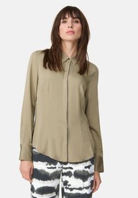 Olivgrüne Bluse mit Kragen, langen Ärmeln und einer Vorderseite mit dekorativen Knöpfen, kombiniert mit gemusterten Schwarz-Weiß-Hosen.