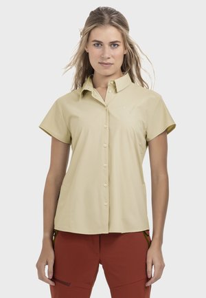 Frau mit langem Haar, die ein beigefarbenes, kurzärmeliges Hemd mit Knöpfen und rostfarbene Hose trägt, steht mit entspannten Armen an den Seiten.