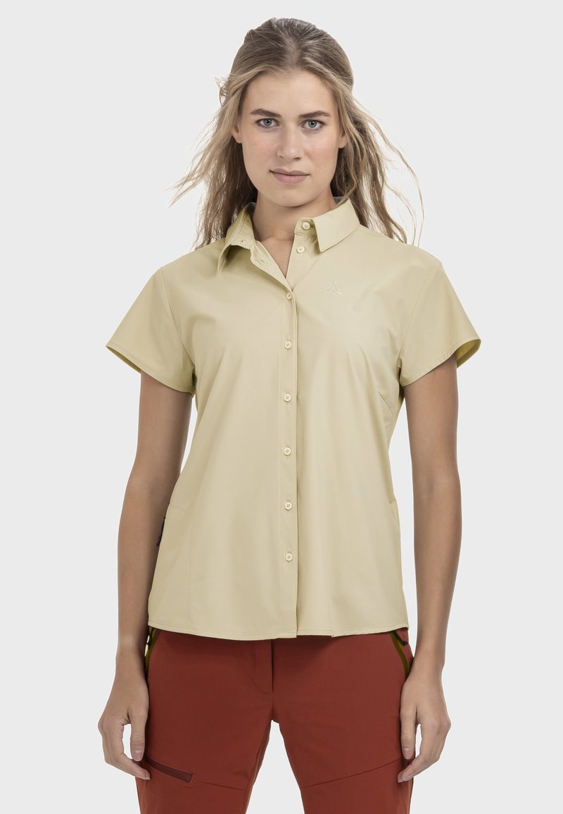 Frau mit langem Haar, die ein beigefarbenes, kurzärmeliges Hemd mit Knöpfen und rostfarbene Hose trägt, steht mit entspannten Armen an den Seiten.