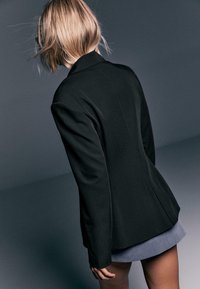 Blazer nero con spalle strutturate e vestibilità sartoriale, abbinato a una gonna grigia. Tessuto liscio con minimalismi e una silhouette pulita.