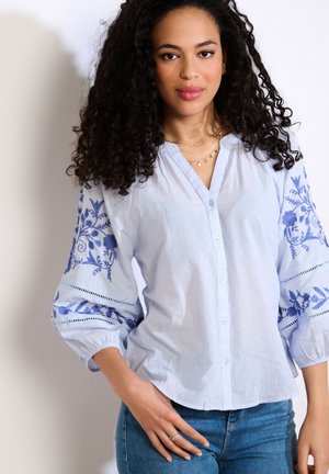 Žena s kučeravými vlasmi, oblečená v svetlomodrej košeli zapínanej na gombíky s modrou kvetinovou výšivkou na nafúknutých rukávoch a modrých džínsach, stojaca pri bielej stene.