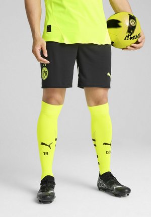 Uniforme da calcio con maglia gialla brillante e calze abbinate fino al ginocchio, pantaloni neri e scarpe da calcio nere con design testurizzato e loghi.