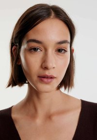 Close-up van een persoon met kort, steil donker haar, een neutrale huidtoon en gekleed in een diep V-hals, geribd bruin kledingstuk. Natuurlijke make-up.
