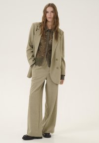 Blazer beige ajustado de corte holgado, combinado con una blusa transparente verde oliva con patrones de encaje floral y pantalones de pierna ancha.