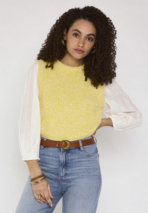 Femme aux cheveux bouclés portant un pull sans manches jaune, un chemisier blanc, un jean bleu, une ceinture marron et des bijoux en or, posant avec la main sur la hanche.