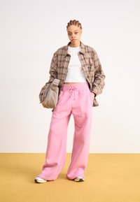 Een bruine geruite overshirt over een witte crop top, gecombineerd met losse roze sweatpants met een trekkoord en het Nike-logo. Grijze schoudertas.