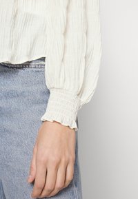 Leichte, strukturierte Bluse mit einem zerknitterten Finish und gerafften, rüschierten Ärmeln, kombiniert mit hellblauen, hochgeschnittenen Jeans.