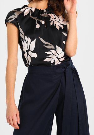 Femme portant un chemisier noir à imprimé floral beige et un pantalon large taille haute bleu marine avec une ceinture nouée à la taille.