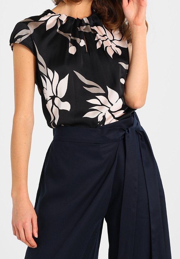 Femme portant un chemisier noir à imprimé floral beige et un pantalon large taille haute bleu marine avec une ceinture nouée à la taille.