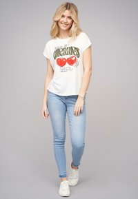 T-shirt blanche avec le texte vert "CIGARIES" et des graphiques de pomme rouge, associée à un jean skinny bleu clair et des baskets blanches. Tissu doux.