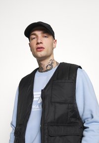 Chaleco acolchado negro con cuatro bolsillos, llevado sobre una sudadera azul claro con un logotipo gráfico. La persona lleva una gorra negra.