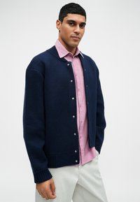 Tumesinine kudumine kootud kardigan ribatud varrukate ja alläärtega, hõbedaste nööpidega, mille all on seljas helesinine vertikaalsete triipudega särk.