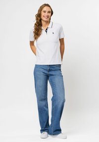 Usmievavá žena s dlhými vlasmi, oblečená v bielej polotričku, modrých džínsach a bielych teniskách, stojaca s rukami za chrbtom.
