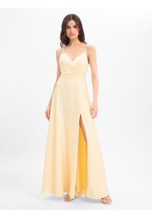 Frau in einem bodenlangen blassgelben Kleid mit Spaghettiträgern und hohem Schlitz, stehend vor einem schlichten weißen Hintergrund.