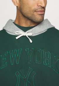 Sudadera verde con capucha gris, con "NUEVA YORK" bordado en letras. Acento de cuerda blanca y tela texturizada.