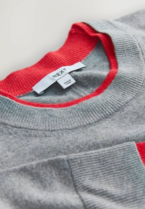 Pelēks džemperis ar sarkanu ribveida apkakli un kabatu, izgatavots no mīksta auduma, ar etiķeti, kurā rakstīts "NEXT" un "MADE IN CHINA".