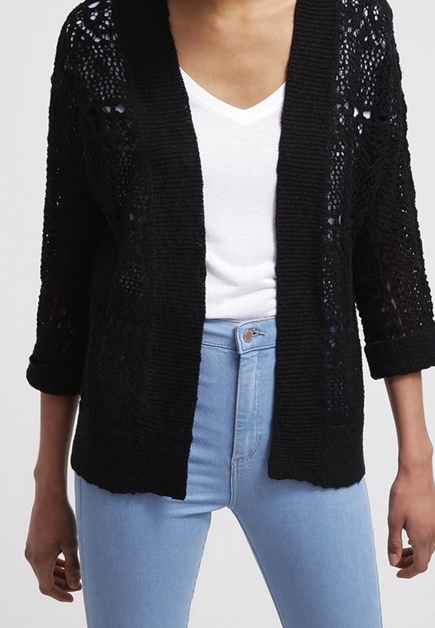 Personne portant un cardigan noir en maille ajourée sur un t-shirt blanc à col en V et un jean taille haute bleu clair.