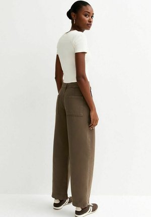 Vrouw staand, gekleed in een wit nauwsluitend kortärmelig shirt, hoge taille wijde bruine broek met achterzakken, en bruine sneakers met witte strepen.