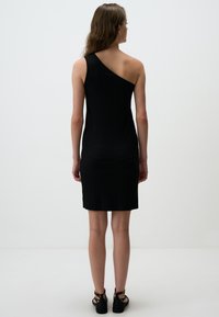Abito nero a coste con design a spalla unica, silhouette aderente e lunghezza fino alla metà della coscia, abbinato a sandali neri con cinturini e texture liscia.