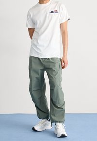 Camiseta blanca de algodón con un logo azul, combinada con pantalones cargo verde claro con bolsillos laterales, y zapatillas deportivas blancas con detalles en negro.