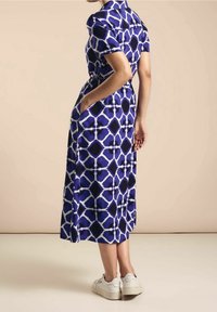 Robe bleue à motifs avec des manches courtes, un col et des poches, présentant un design géométrique dans des nuances sombres et claires. Portée avec des baskets blanches.