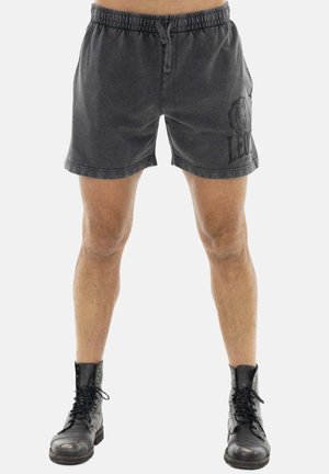 Uomo che indossa pantaloncini neri con coulisse e logo di leone sul lato sinistro, abbinati a stivali neri usurati con lacci, in piedi su sfondo bianco.