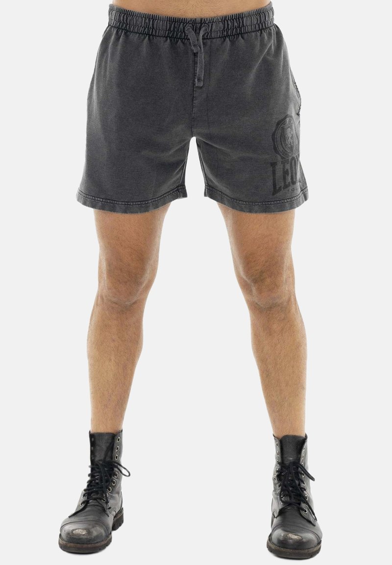 Homme portant un short noir à cordon avec un logo de lion sur le côté gauche, associé à des bottes noires usées à lacets, debout sur un fond blanc.
