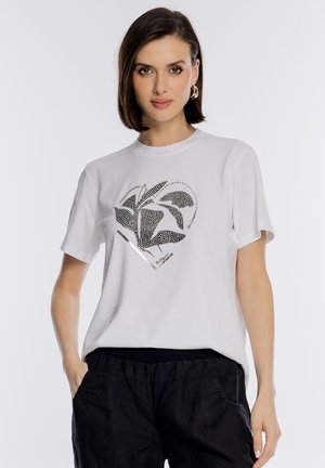 Kobieta z krótkimi brązowymi włosami, nosząca złote kolczyki koła, białą koszulkę z czarnym wzorem liści oraz czarne spodnie, stoi z rękami w kieszeniach.