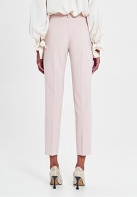 Pantalons taillés rose clair avec une texture lisse, présentant une coupe slim et des jambes fuselées, associés à des chaussures à talons hauts en sequins.