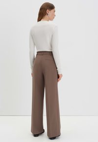 Braune weite Hosen mit glatter Textur und Gesäßtaschen, kombiniert mit einem taillierten weißen Langarmoberteil, von hinten betrachtet.