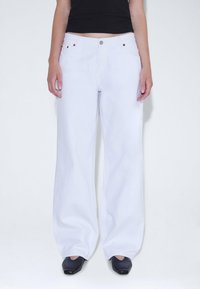 Jeans de pierna ancha blancos con cintura alta, hechos de denim. Presentan herrajes en tono dorado, cierre de botón y bolsillos laterales.
