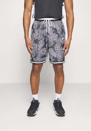 Man draagt zwarte sportshorts met wit abstract patroon, zwart tanktop, witte sokken en zwarte sneakers, staand tegen een effen achtergrond.