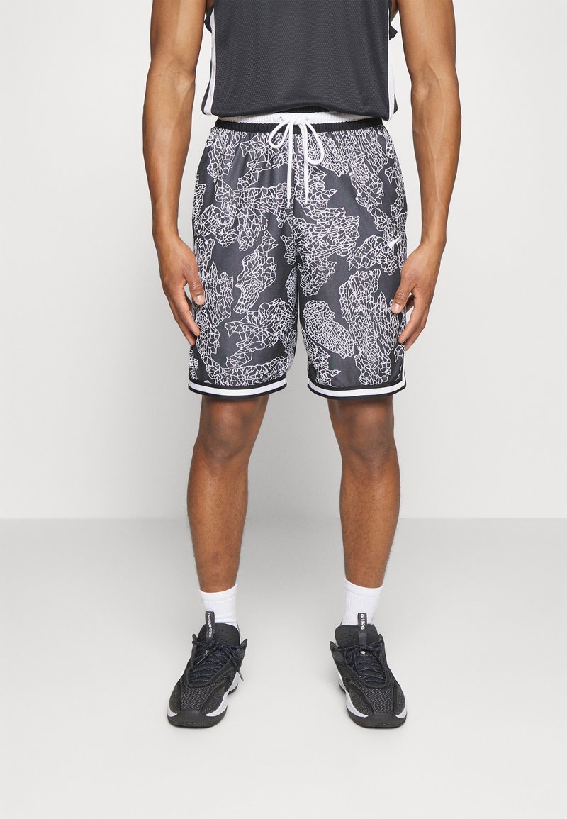 Homme portant un short de sport noir avec un motif abstrait blanc, un débardeur noir, des chaussettes blanches et des baskets noires, debout devant un fond uni.