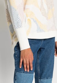 Sudadera en blanco roto con patrones abstractos multicolores en rosa, amarillo, azul y marrón. Puños acanalados; combinada con pantalones de mezclilla azul.