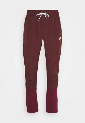 Maroon Nike joggingbroek met elastische tailleband, witte trekkoord, wit Nike-logo op de linker dij en donkerdere maroon panelen aan de onderkant van de benen.