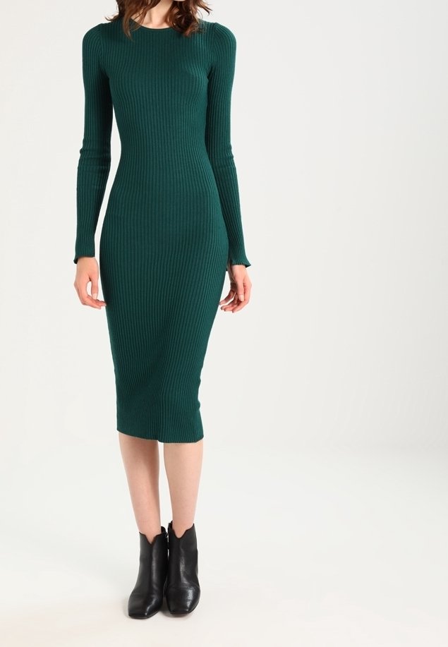 Femme portant une robe midi ajustée, à manches longues et côtelée de couleur vert foncé, associée à des bottines noires sur fond blanc.