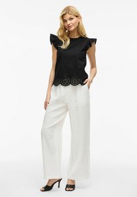 Blusa nera con maniche a volant e orlo con taglio laser, abbinata a pantaloni bianchi a gamba larga e décolleté neri open-toe.