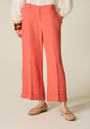 Kobieta ubrana w koralowe, szerokie, skrócone spodnie, beżowe mokasyny, białą bluzkę i masywne bransoletki na lewej ręce, stojąca na beżowej podłodze.