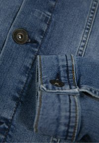 Szczegół kurtki dżinsowej z niebieskiego materiału, widoczne szwy i czarny guzik na mankiecie. Miękka tekstura z klasycznym wzornictwem.