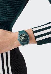 Grön silikonklocka med en rund urtavla som har en minimalistisk design och randigt mönster. Bärs på ett handled med en svart- och vitt randig ärm.