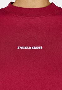 Piros pamut kerek nyakú póló bordázott nyakkivágással és fehér hímzett "PEGADOR" logóval az elején. Sima textúra, minimalista kialakítás.