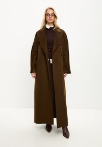 Langer brauner Wollmantel mit doppelter Knopfreihe, großen Revers und Frontknöpfen. Getragen über einem dunklen Pullover, kombiniert mit braunen Absatzstiefeln.